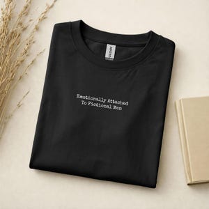 Może przedstawiać: Czarny t-shirt z okrągłym dekoltem z białym napisem "Emotionally Attached To Fictional Men". Koszulka jest złożona na jasnej powierzchni, z książką i suszonymi roślinami w tle. Widoczna jest metka Gildan.