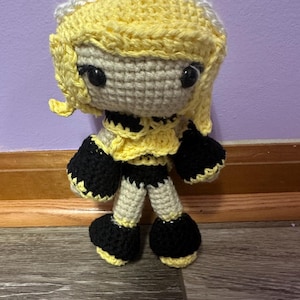 May include: A handmade crochet doll with a yellow and black outfit, yellow hair, and black eyes. The doll has a white headpiece and is standing on a wooden surface. The doll's outfit includes a yellow top with black accents and black shorts.
