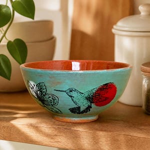 Pode incluir: Uma tigela de cerâmica redonda com exterior turquesa e interior laranja quente. O exterior apresenta uma ilustração de linhas pretas de um beija-flor com uma asa vermelha brilhante e uma flor, sobre um acabamento liso e esmaltado.