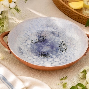 May include: A round, shallow terracotta bowl with two side handles. The interior features a light blue and white mottled glaze with black line drawings of a seahorse, a crab, and a seashell centered on the base of the dish.