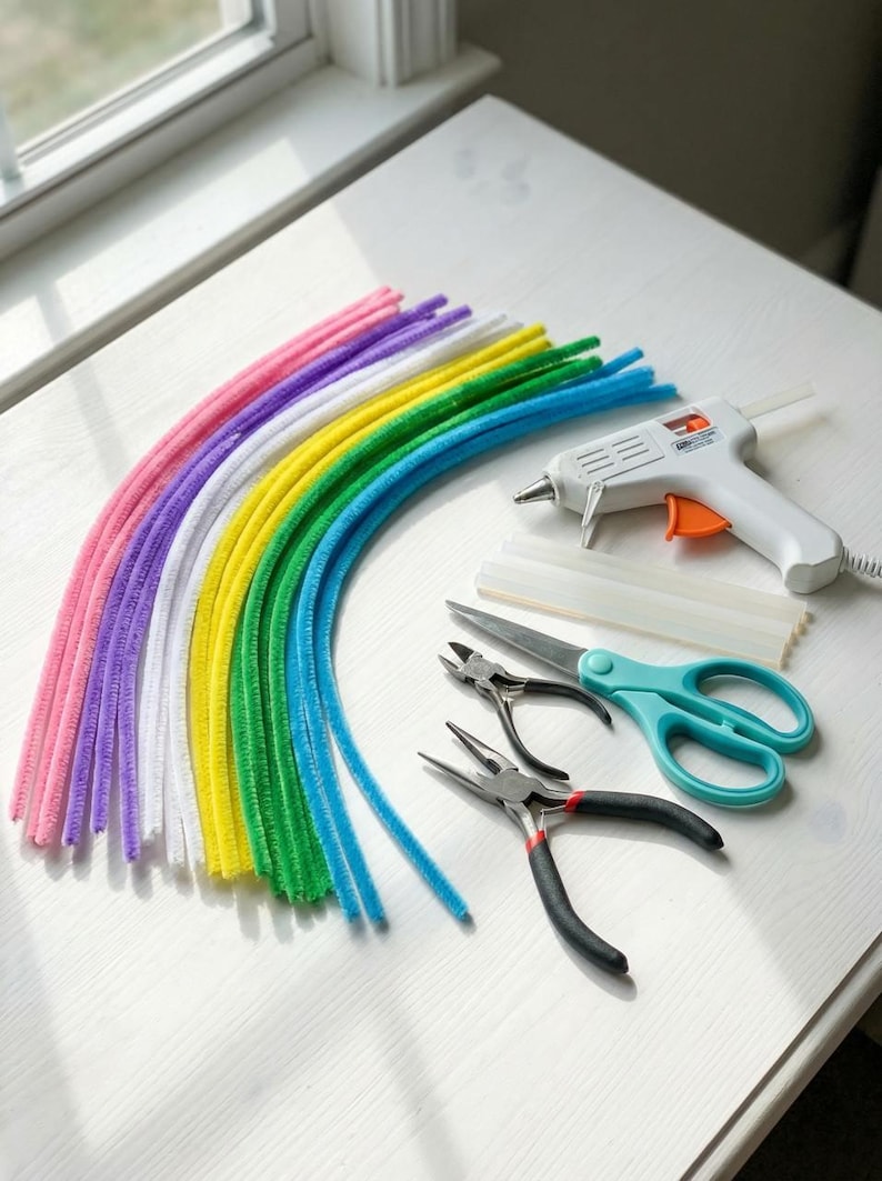 Pode incluir: Uma cole&ccedil;&atilde;o de materiais de artesanato coloridos, incluindo um arco-&iacute;ris de limpadores de cachimbo, uma pistola de cola com bast&otilde;es de cola, tesouras e dois pares de alicates. As ferramentas est&atilde;o dispostas sobre uma superf&iacute;cie branca perto de uma janela.