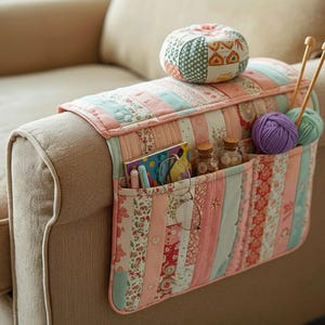 Puede incluir: Organizador de reposabrazos acolchado en colores pastel, con múltiples bolsillos para guardar cosas. El organizador está lleno de materiales de manualidades, como hilo, agujas de tejer y pequeños frascos de vidrio. Encima hay un pequeño cojín redondo de patchwork.
