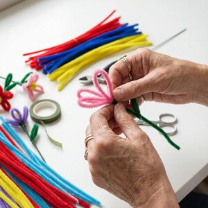Pode incluir: Uma pessoa est&aacute; a fazer uma flor rosa com arame de chenille e haste verde. H&aacute; feixes de arames de chenille coloridos em vermelho, azul, amarelo e roxo espalhados. Tesouras, cortadores de arame e fita floral tamb&eacute;m est&atilde;o presentes, sugerindo um projeto de artesanato.