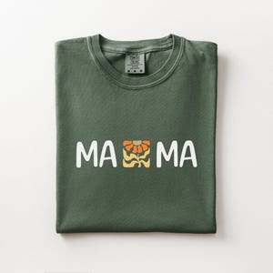 Folded moss Comfort Colors shirt featuring white MAMA lettering with a retro floral icon in warm orange and golden tones.