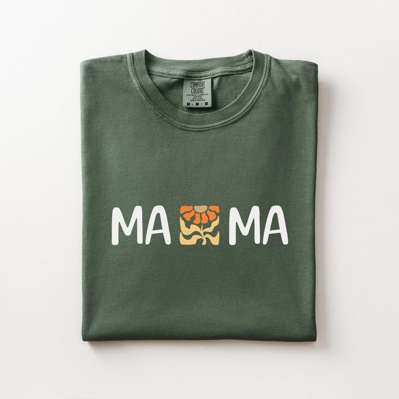 Folded moss Comfort Colors shirt featuring white MAMA lettering with a retro floral icon in warm orange and golden tones.