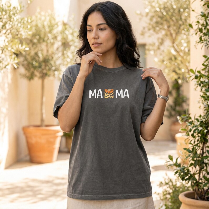 Woman wearing a pepper Comfort Colors mama shirt with white MAMA lettering and a retro floral icon in warm orange and golden tones.
