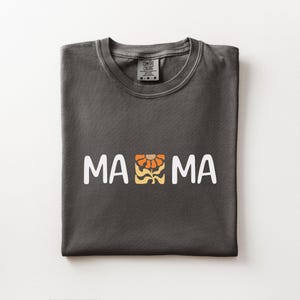 Folded pepper Comfort Colors shirt featuring white MAMA lettering with a retro floral icon in warm orange and golden tones.