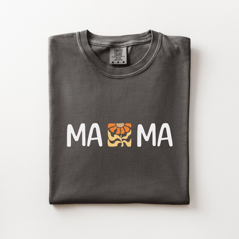 Folded pepper Comfort Colors shirt featuring white MAMA lettering with a retro floral icon in warm orange and golden tones.