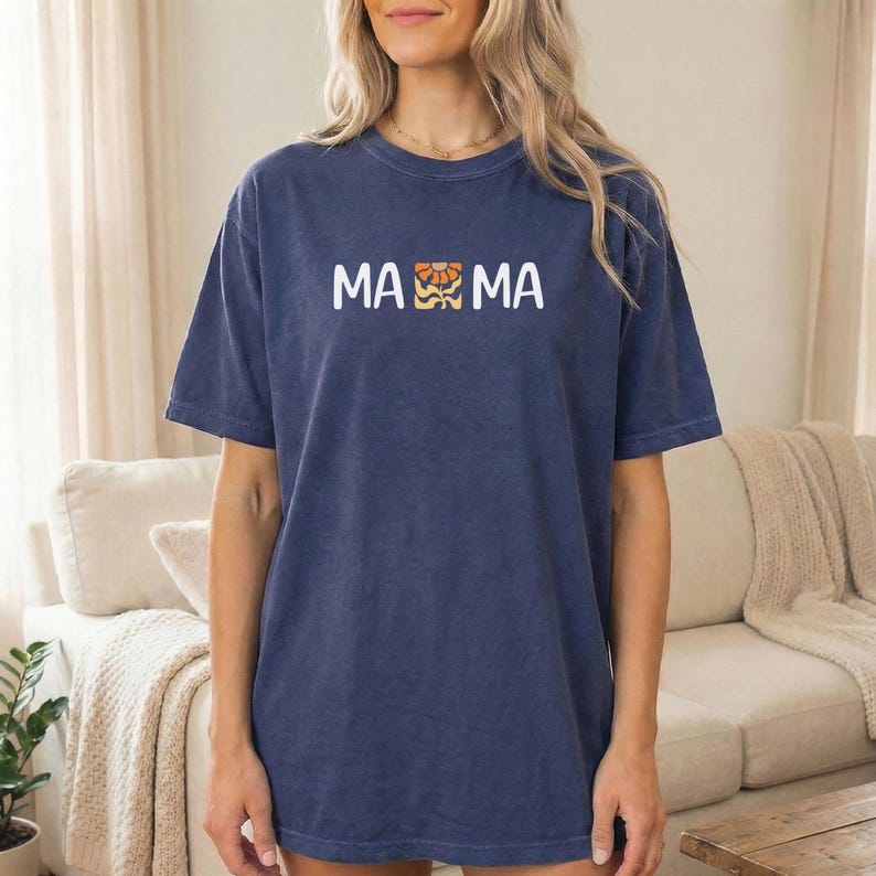 Woman wearing a navy Comfort Colors mama shirt with white MAMA lettering and a retro floral icon in warm orange and golden tones.