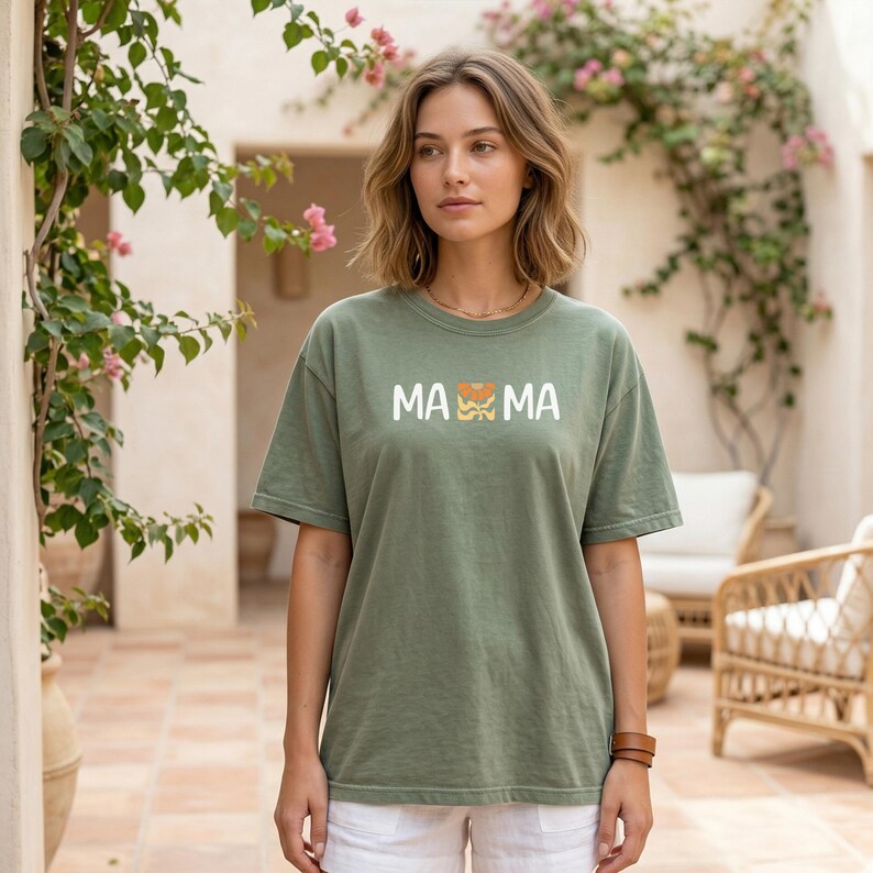 Woman wearing a moss Comfort Colors mama shirt with white MAMA lettering and a retro floral icon in warm orange and golden tones.
