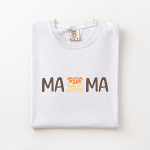 Folded white Comfort Colors shirt featuring brown MAMA lettering with a retro floral icon in orange, beige, and golden tones.