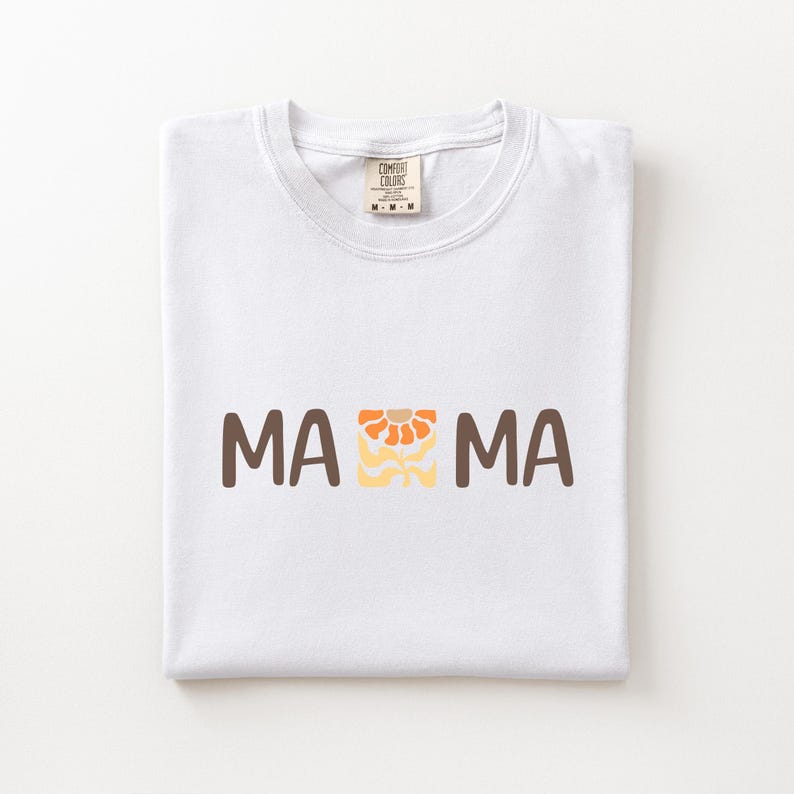 Folded white Comfort Colors shirt featuring brown MAMA lettering with a retro floral icon in orange, beige, and golden tones.