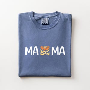 Folded blue jean Comfort Colors shirt featuring white MAMA lettering with a retro floral icon in warm orange and golden tones.