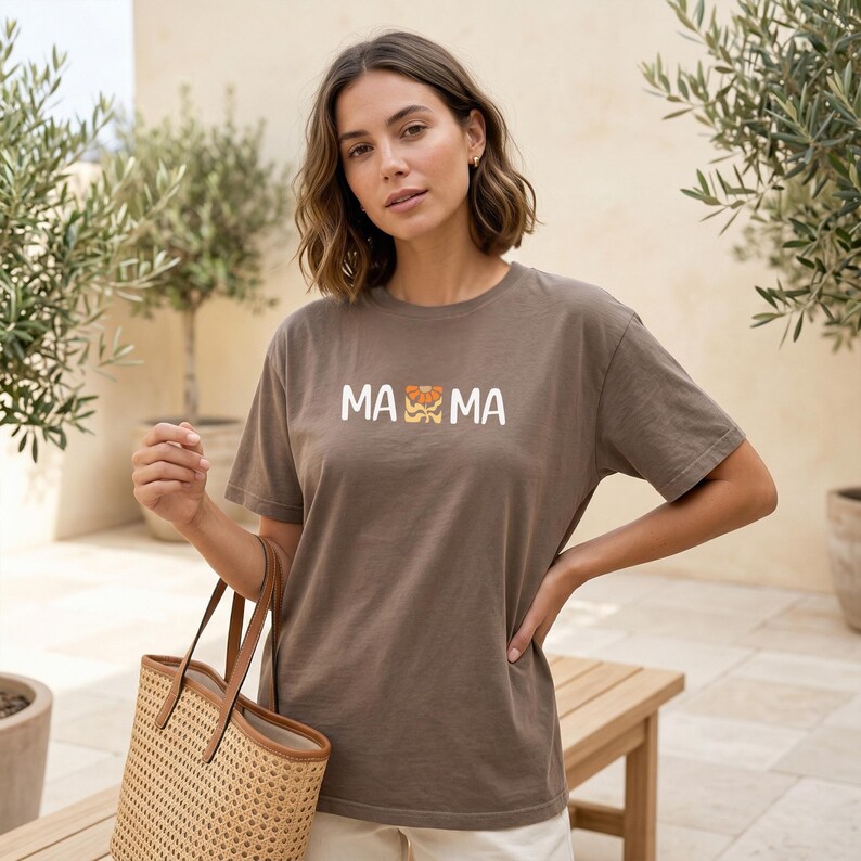Woman wearing an espresso Comfort Colors mama shirt with white MAMA lettering and a retro floral icon in warm orange and golden tones.