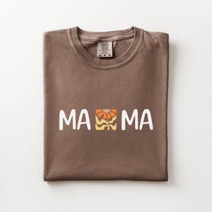 Folded espresso Comfort Colors shirt featuring white MAMA lettering with a retro floral icon in warm orange and golden tones.