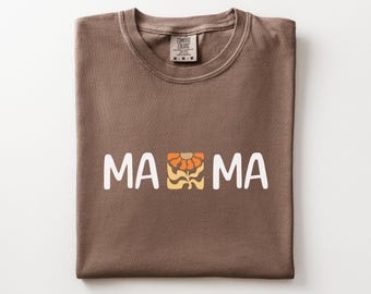May include: A brown t-shirt with the word "MA MA" in white block letters. A floral design in orange, yellow, and brown is between the words. The shirt is folded neatly and has a Comfort Colors tag.