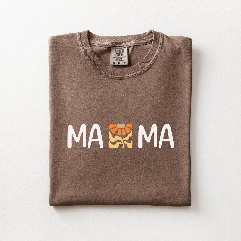 Folded espresso Comfort Colors shirt featuring white MAMA lettering with a retro floral icon in warm orange and golden tones.