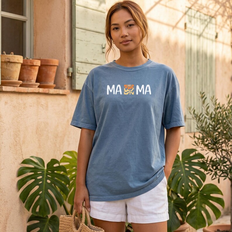 Woman wearing a blue jean Comfort Colors mama shirt with white MAMA lettering and a retro floral icon in warm orange and golden tones.