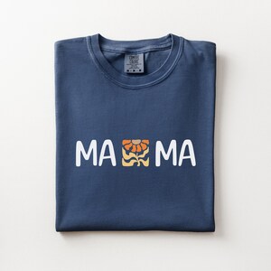 Folded navy Comfort Colors shirt featuring white MAMA lettering with a retro floral icon in warm orange and golden tones.