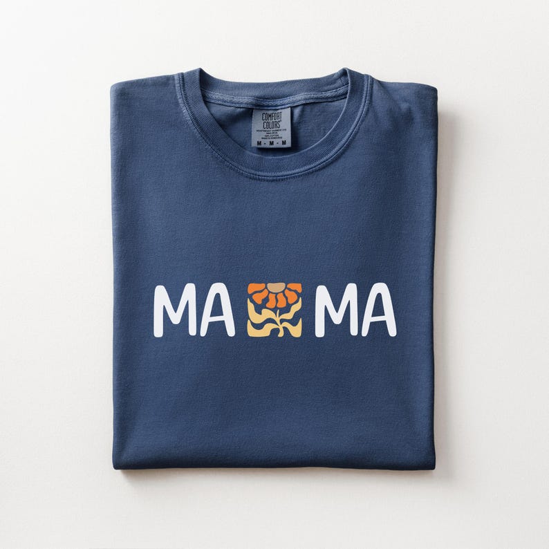 Folded navy Comfort Colors shirt featuring white MAMA lettering with a retro floral icon in warm orange and golden tones.