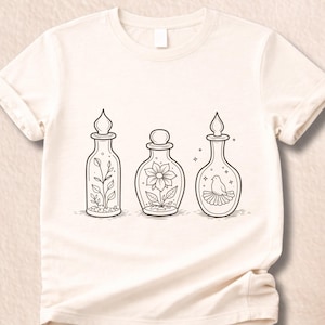 May include: Cream-colored t-shirt featuring three decorative bottles with black line art. The bottles contain a plant, a flower, and a bird. The shirt has short sleeves and a crew neck.