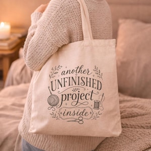 May include: A beige canvas tote bag featuring black printed text that reads another UNFINISHED project inside, accompanied by illustrations of a ball of yarn, a spool of thread, and a pair of scissors, all surrounded by decorative leaf accents.
