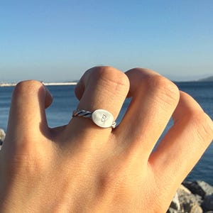 Puede incluir: Un anillo de tono plateado con una banda trenzada y una pieza central lisa de forma ovalada. La cara ovalada está grabada con la letra mayúscula B. El anillo tiene un acabado metálico y un diseño texturizado similar a una cuerda.