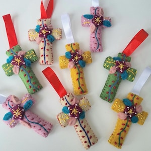 May include: A selection of handmade felt crosses in shades of pink, green, and yellow. Each cross features embroidered floral designs and a ribbon for hanging. These decorative crosses are a charming, handcrafted item.