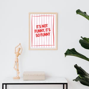 May include: A framed print with the text "IT'S NOT FUNNY, IT'S SO FUNNY" in red on a pink and white striped background. The frame is light brown. A wooden artist's mannequin and a beige box sit on a white table.