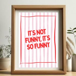 May include: A framed print featuring the phrase "IT'S NOT FUNNY, IT'S SO FUNNY" in red, on a backdrop of pink and white vertical stripes. The frame is made of light brown wood.