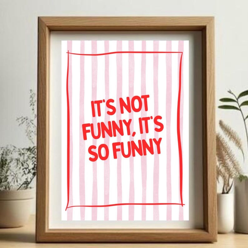 May include: A framed print featuring the phrase "IT'S NOT FUNNY, IT'S SO FUNNY" in red, on a backdrop of pink and white vertical stripes. The frame is made of light brown wood.