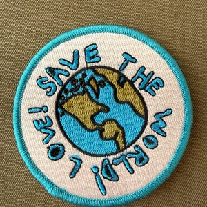 May include: Embroidered patch with a light blue border and the words "SAVE THE WORLD! LOVE!" in light blue lettering. The patch features a globe design with blue oceans and green and yellow continents.