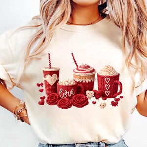 Könnte beinhalten: Cremefarbenes Sweatshirt mit einem Valentinstagsdesign. Die Grafik zeigt rote Becher, Tassen und Rosen mit dem Wort "love" und Herzakzenten. Das Design hat eine gestrickte Optik.