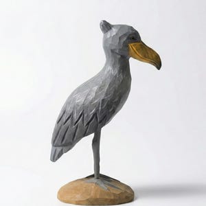 Puede incluir: Escultura de madera tallada a mano de un picozapato. El ave es gris con un pico amarillo dorado y se apoya sobre una base marr&oacute;n claro. La escultura tiene un aspecto texturizado y tallado, y el fondo es blanco.