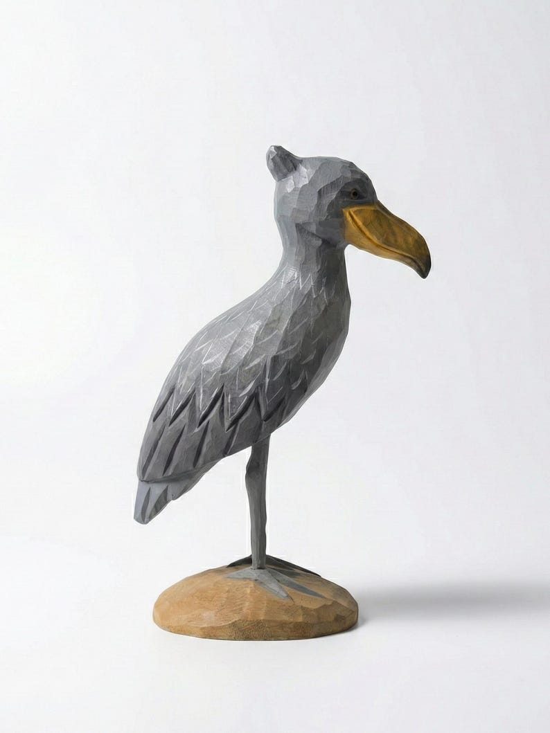 Puede incluir: Escultura de madera tallada a mano de un picozapato. El ave es gris con un pico amarillo dorado y se apoya sobre una base marr&oacute;n claro. La escultura tiene un aspecto texturizado y tallado, y el fondo es blanco.