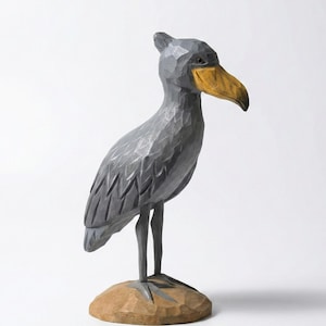 Puede incluir: Escultura de madera tallada a mano de un ave picozapato. El ave es principalmente gris con un pico amarillo y se apoya sobre una peque&ntilde;a base de madera marr&oacute;n. La escultura tiene un aspecto texturizado y facetado, sobre un fondo blanco.
