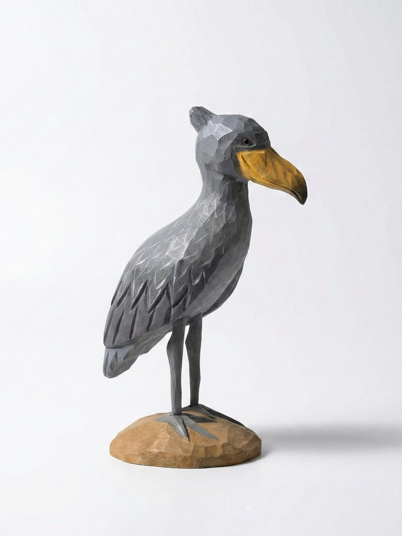 Puede incluir: Escultura de madera tallada a mano de un ave picozapato. El ave es principalmente gris con un pico amarillo y se apoya sobre una peque&ntilde;a base de madera marr&oacute;n. La escultura tiene un aspecto texturizado y facetado, sobre un fondo blanco.