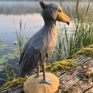 Puede incluir: Escultura de madera tallada a mano de un picozapato. El ave es gris con un gran pico amarillo y se apoya sobre una base marr&oacute;n claro tallada. La escultura se encuentra sobre un fondo borroso de un lago y vegetaci&oacute;n.