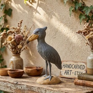 Puede incluir: Una escultura de madera tallada a mano de un picozapato, pintada de gris con un pico amarillo, se alza sobre una base de madera tallada. La escultura se exhibe en una estanter&iacute;a de madera r&uacute;stica con un letrero que dice "HANDCARVED ART-ETSY".
