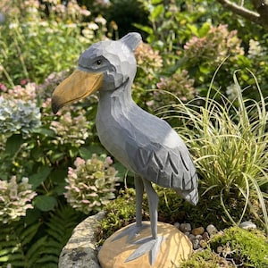 Puede incluir: Una escultura de madera gris de un picozapato con un pico grande de color amarillo-marr&oacute;n. El ave se alza sobre una base de madera tallada con musgo y peque&ntilde;as piedras, colocada en una jardinera de piedra. El fondo presenta exuberante vegetaci&oacute;n y flores.