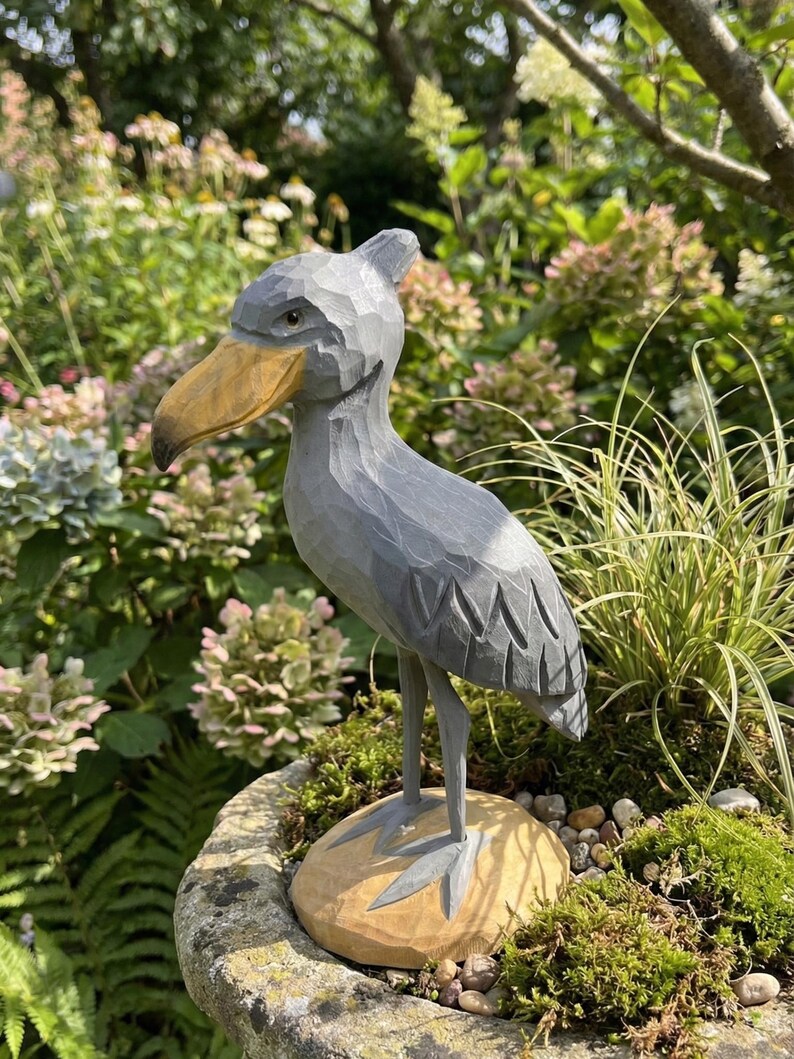 Puede incluir: Una escultura de madera gris de un picozapato con un pico grande de color amarillo-marr&oacute;n. El ave se alza sobre una base de madera tallada con musgo y peque&ntilde;as piedras, colocada en una jardinera de piedra. El fondo presenta exuberante vegetaci&oacute;n y flores.