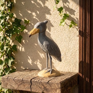 Puede incluir: Una escultura de madera gris de un ave picozapato con pico amarillo se encuentra sobre una estanter&iacute;a de madera. La escultura presenta detalles de plumas talladas y una superficie texturizada. La estanter&iacute;a es de madera en bruto, y el fondo presenta una pared texturizada y vegetaci&oacute;n.