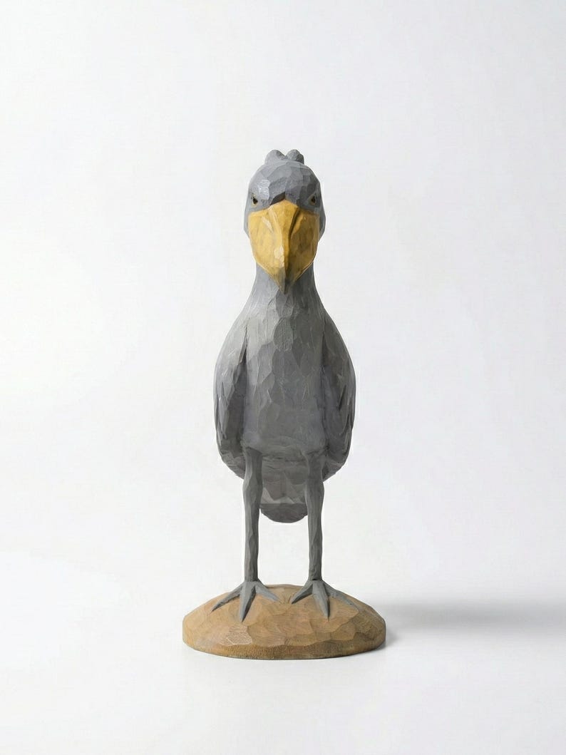 Puede incluir: Una escultura de madera gris de un ave picozapato con un gran pico amarillo. El ave se alza sobre una peque&ntilde;a base tallada de color marr&oacute;n claro. La escultura tiene un aspecto texturizado y tallado, sobre un fondo blanco.