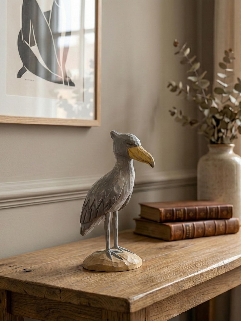 Puede incluir: Escultura de madera tallada a mano de un ave picozapato. El ave es gris con un pico amarillo y se apoya sobre una base marr&oacute;n claro. La escultura se exhibe sobre una mesa de madera, con dos libros encuadernados en cuero y un jarr&oacute;n al fondo.
