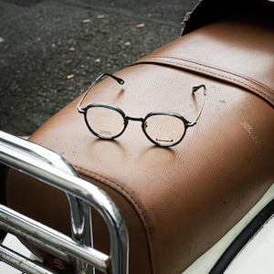 Peut inclure: Des lunettes rondes à monture noire, de design classique, reposent sur une selle en cuir marron. Les lunettes ont des branches fines en métal et des verres clairs. L'arrière-plan comprend un porte-bagages chromé et une partie d'un véhicule blanc.