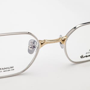 Peut inclure: Gros plan de lunettes avec une monture argentée et des détails dorés sur le pont. Les verres sont clairs, et la monture porte les mentions "TITANIUM" et "BROOKLYN". Les lunettes ont un design géométrique moderne.