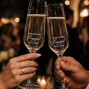 Może przedstawiać: Dwie przezroczyste szklane kieliszki do szampana wypełnione musującym płynem. Każdy kieliszek jest wygrawerowany napisem James & Beth, Engaged oraz datą 02.07.2026. Wysokie, smukłe nóżki i okrągłe podstawy są uniesione w geście toastu.