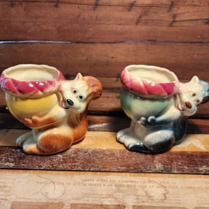 May include: Two ceramic squirrel figurines holding a pot. The squirrel on the left is brown and the squirrel on the right is blue. Both squirrels have red and white accents on their pot.