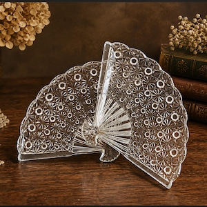 May include: Two clear, fan-shaped glass dishes with intricate starburst and circle patterns. The dishes are displayed on a dark wooden surface, with dried flowers and antique books in the background. The glass has a scalloped edge.
