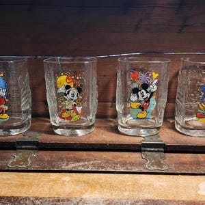 May include: Four clear drinking glasses with different colorful Mickey Mouse designs. Each glass features a unique illustration of Mickey in various outfits and poses. The glasses are displayed on a wooden surface.